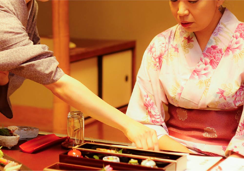 A woman in a colorful pink floral yukata sitting, being served her meal by ryokan staff